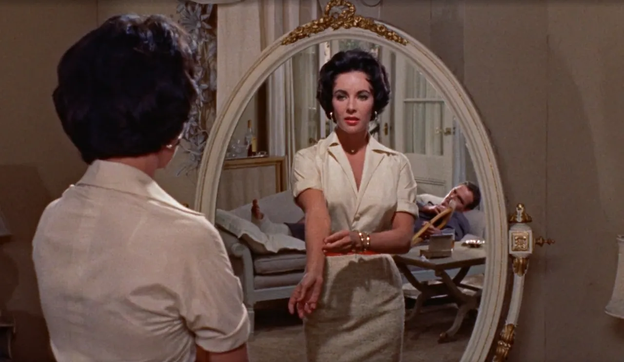 Maggie Pollitt (Elizabeth Taylor) looks into an oval vanity mirror while Brick Pollitt (Paul Newman) reclines behind her with a cast on his leg and a drink in his hand.